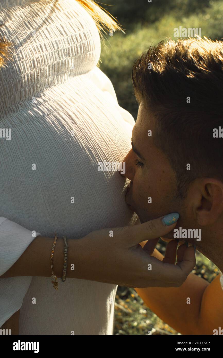 Primer plano de marido hispano besando a su avanzada mujer embarazada vientre con ojos cerrados en un día soleado al aire libre. Foto de stock