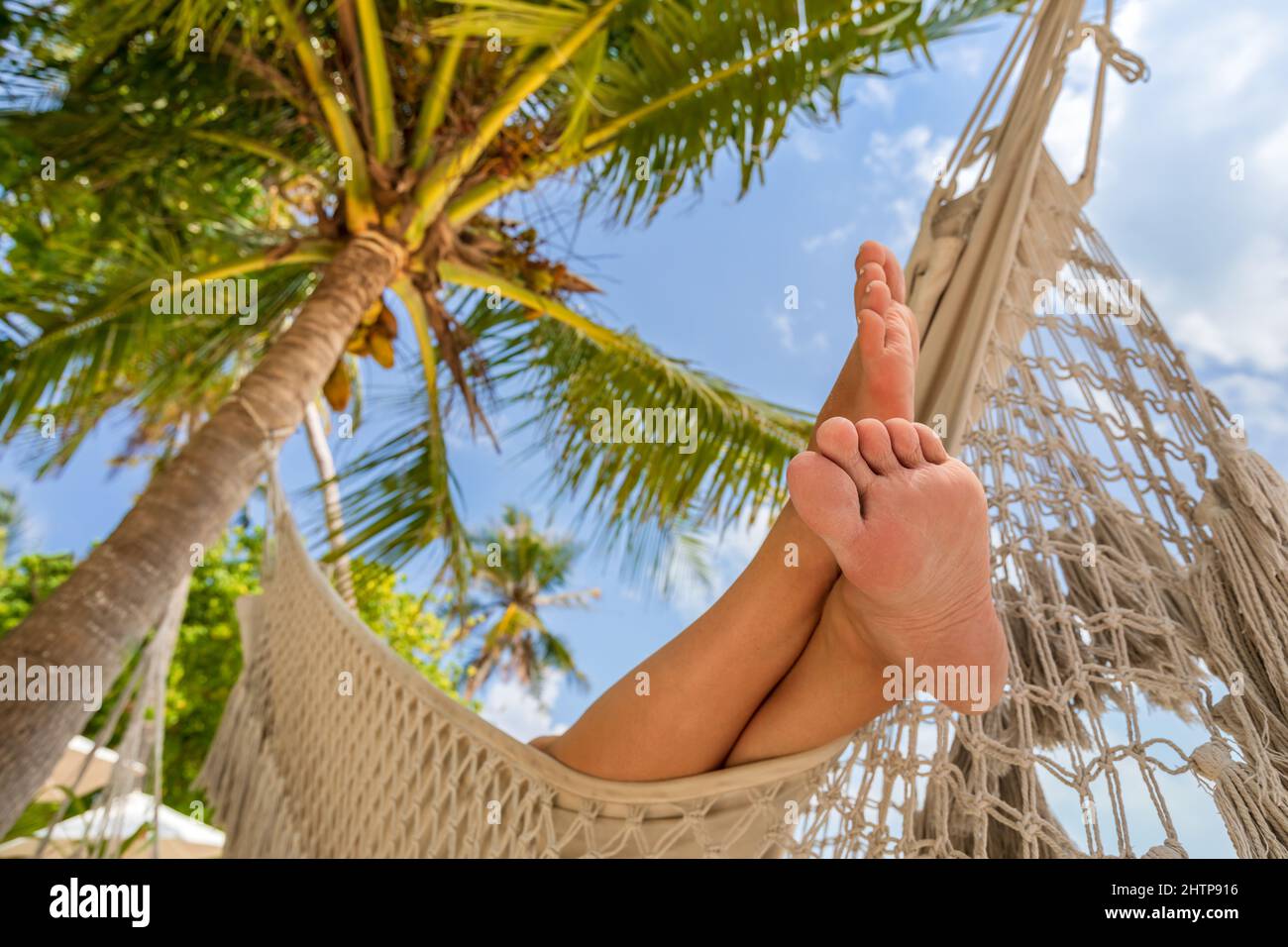 Vacaciones relajantes en la playa con pies de mujer en una hamaca entre palmeras de coco. Exótico complejo hotelero en una isla tropical. Soleado día cálido con cielo azul. Foto de stock