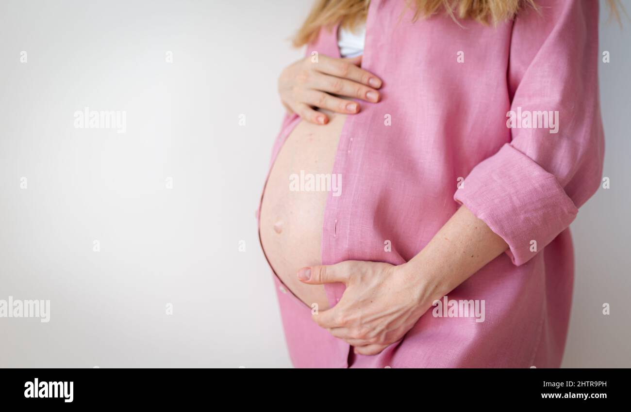 mujer embarazada en una camisa rosa se levanta sobre un fondo blanco y sostiene sus manos sobre su estómago, esperanza, nuevo concepto de vida Foto de stock