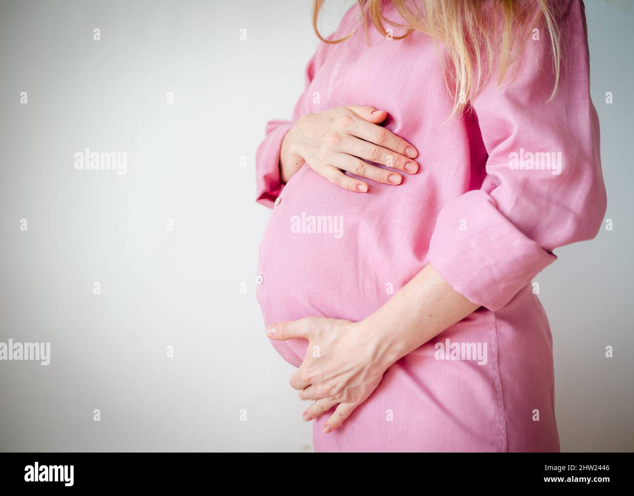 mujer embarazada en una camisa rosa sostiene sus manos sobre su estómago sobre un fondo blanco Foto de stock