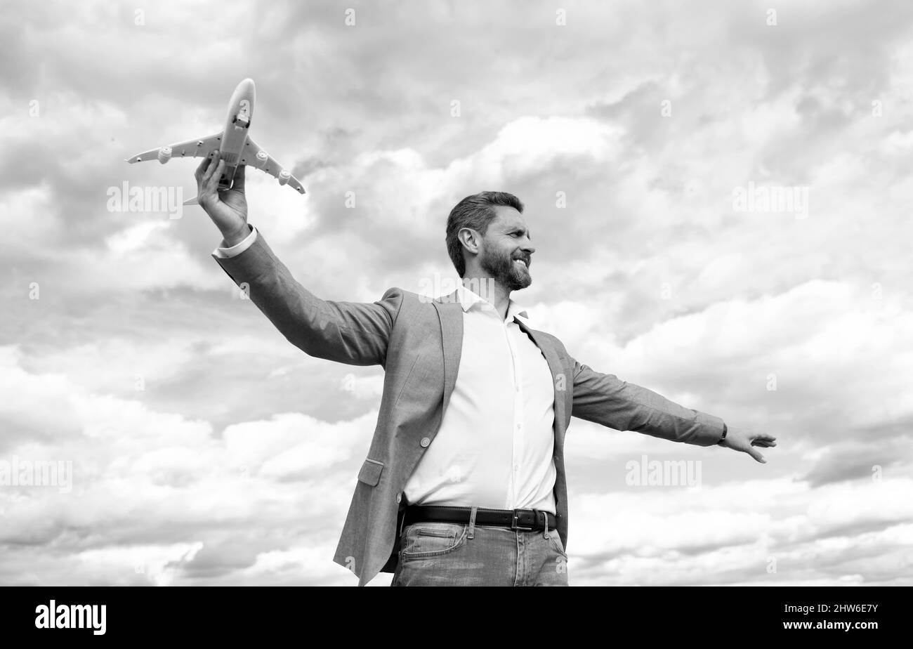 un hombre de negocios con chaqueta, un hombre feliz y guapo, sostiene el avión de juguete sobre el fondo del cielo, viaje de negocios Foto de stock