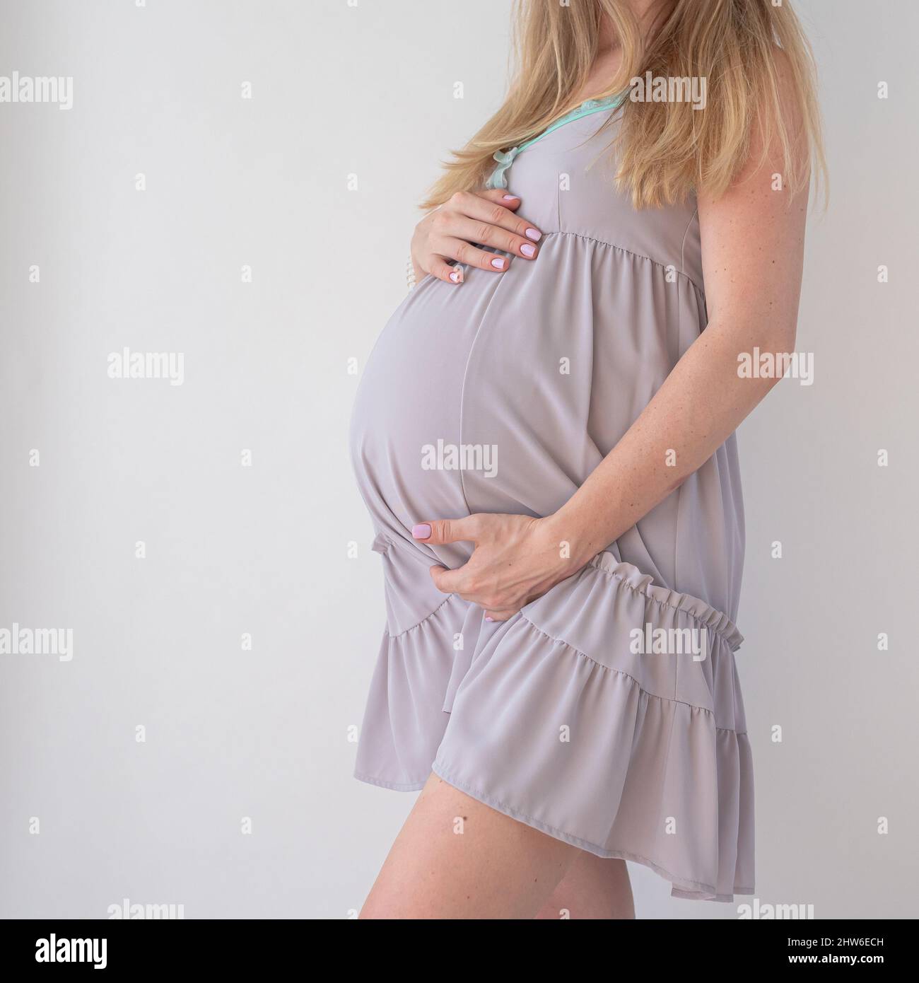 una mujer embarazada con una bata de noche de lila pálida se pone de pie sobre un fondo blanco y sostiene las manos sobre su estómago Foto de stock
