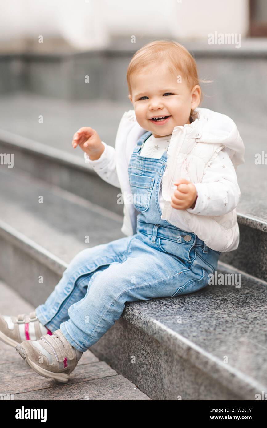 Lindo bebé sonriente niña de 1-2 años de edad llevar pantalones denim sport  y la parte superior blanca en la calle de la ciudad al aire libre. Mirando  la cámara. Infancia. Primavera