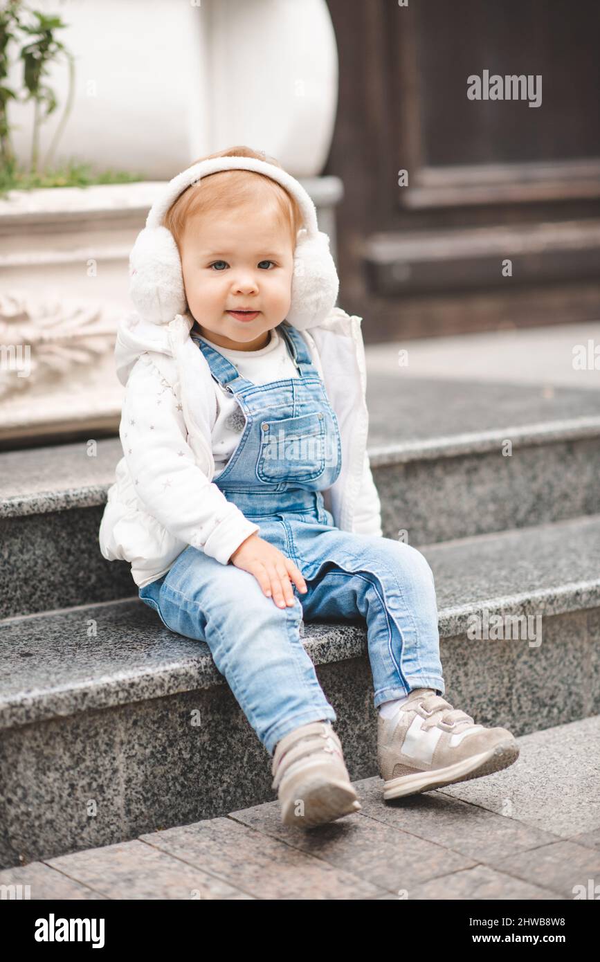 Lindo bebé sonriente niña de 1-2 años de edad llevar pantalones denim sport  y la parte superior blanca en la calle de la ciudad al aire libre. Mirando  la cámara. Infancia. Primavera
