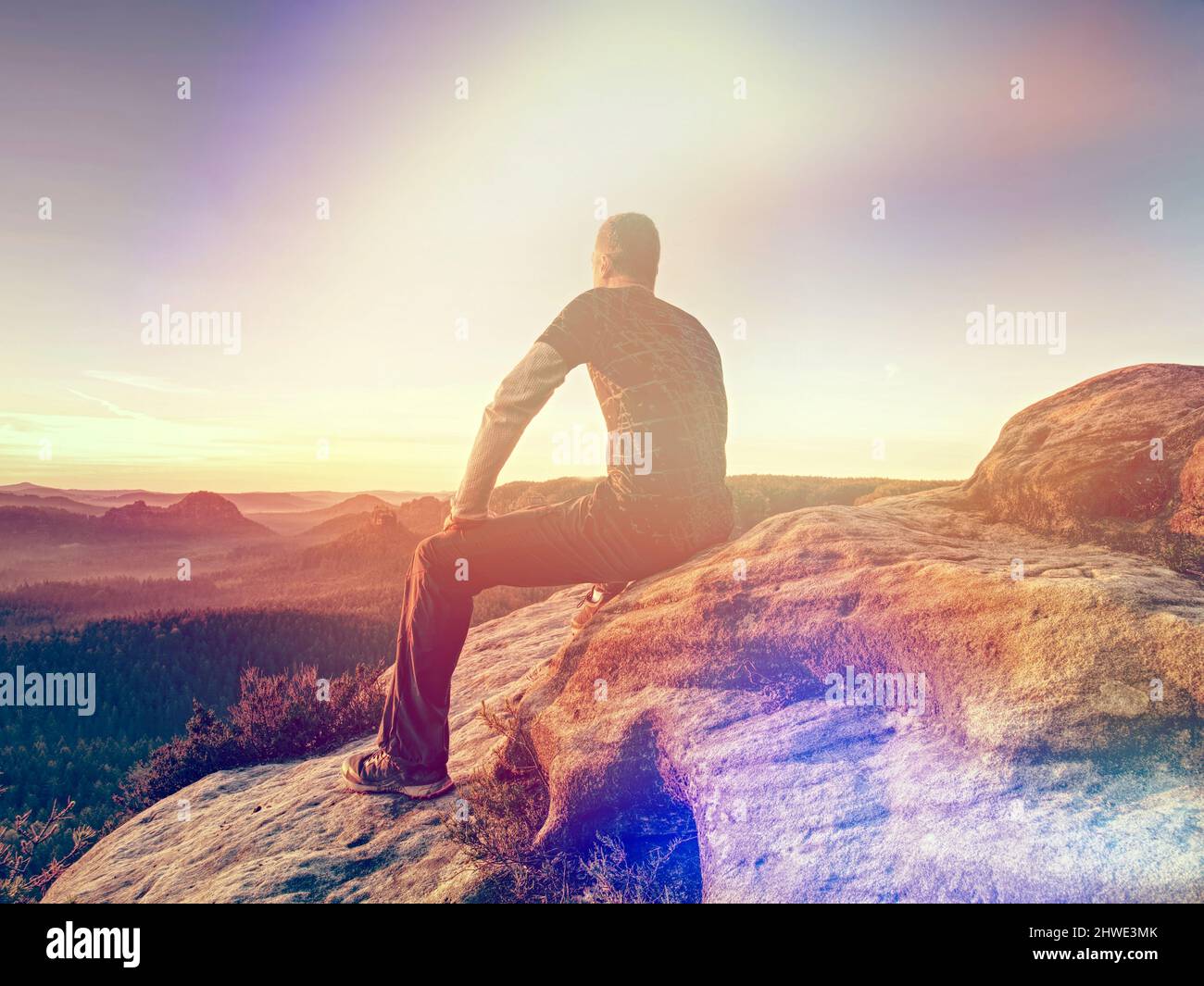 El turista se sienta en una piedra en el borde del acantilado con vistas a las llanuras debajo en el borde brumoso del horizonte. Vista trasera. Iluminación abstracta, llamaradas coloridas. Foto de stock