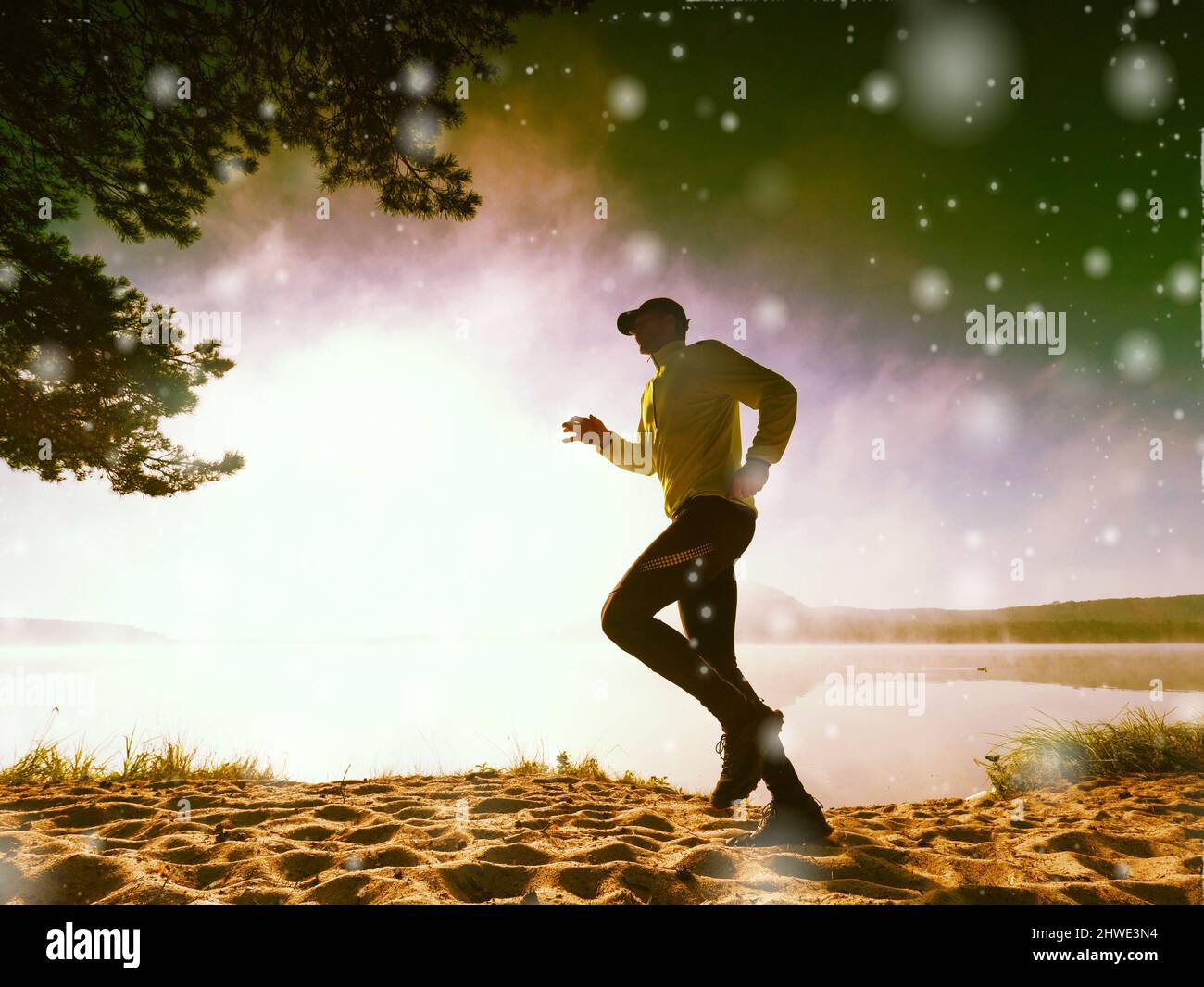 Congele el movimiento del hombre corriendo en la playa de arena en el lago. Chellar copos de nieve de nuevo sol bajo. Iluminación abstracta, llamaradas coloridas. Foto de stock