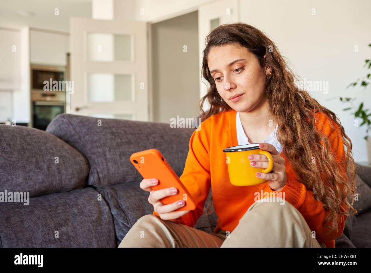 Mujer joven vestida en color naranja con el teléfono inteligente en el sofá Foto de stock