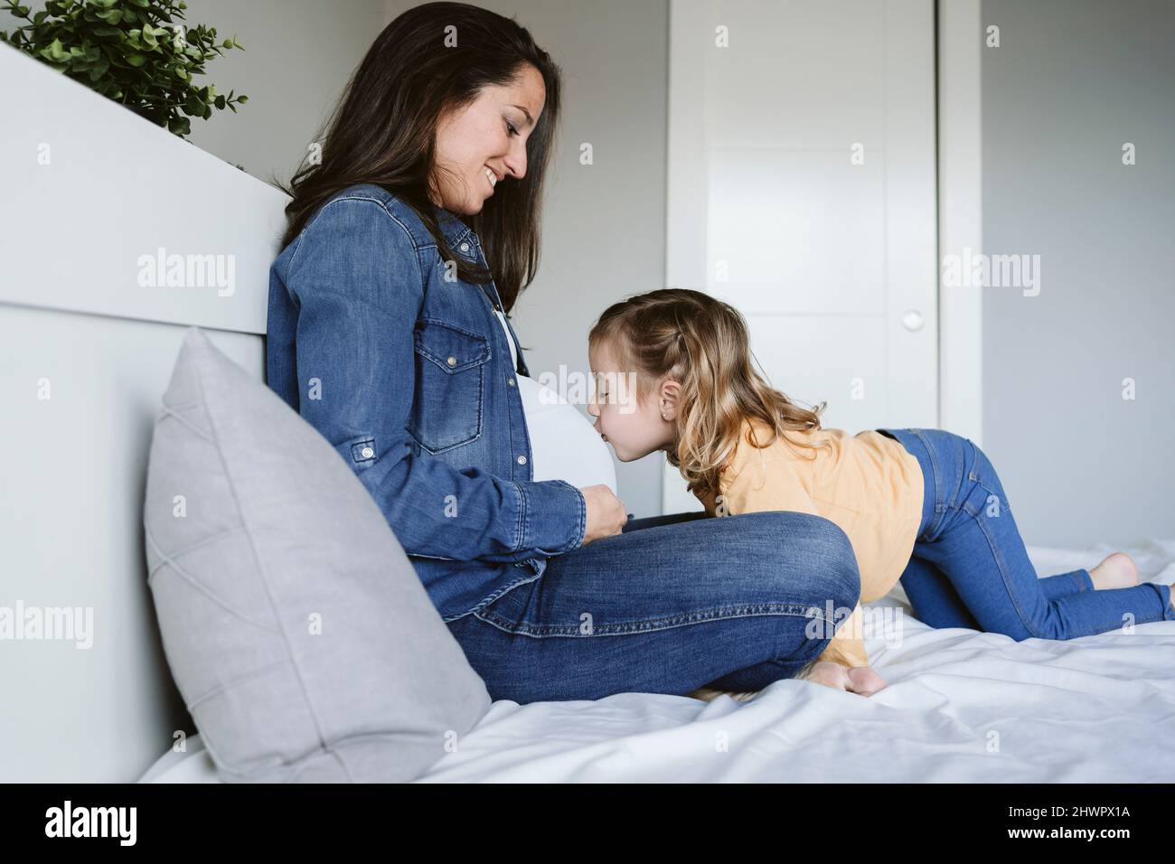 Chica con los ojos cerrados besando el vientre de la madre embarazada en la cama en casa Foto de stock