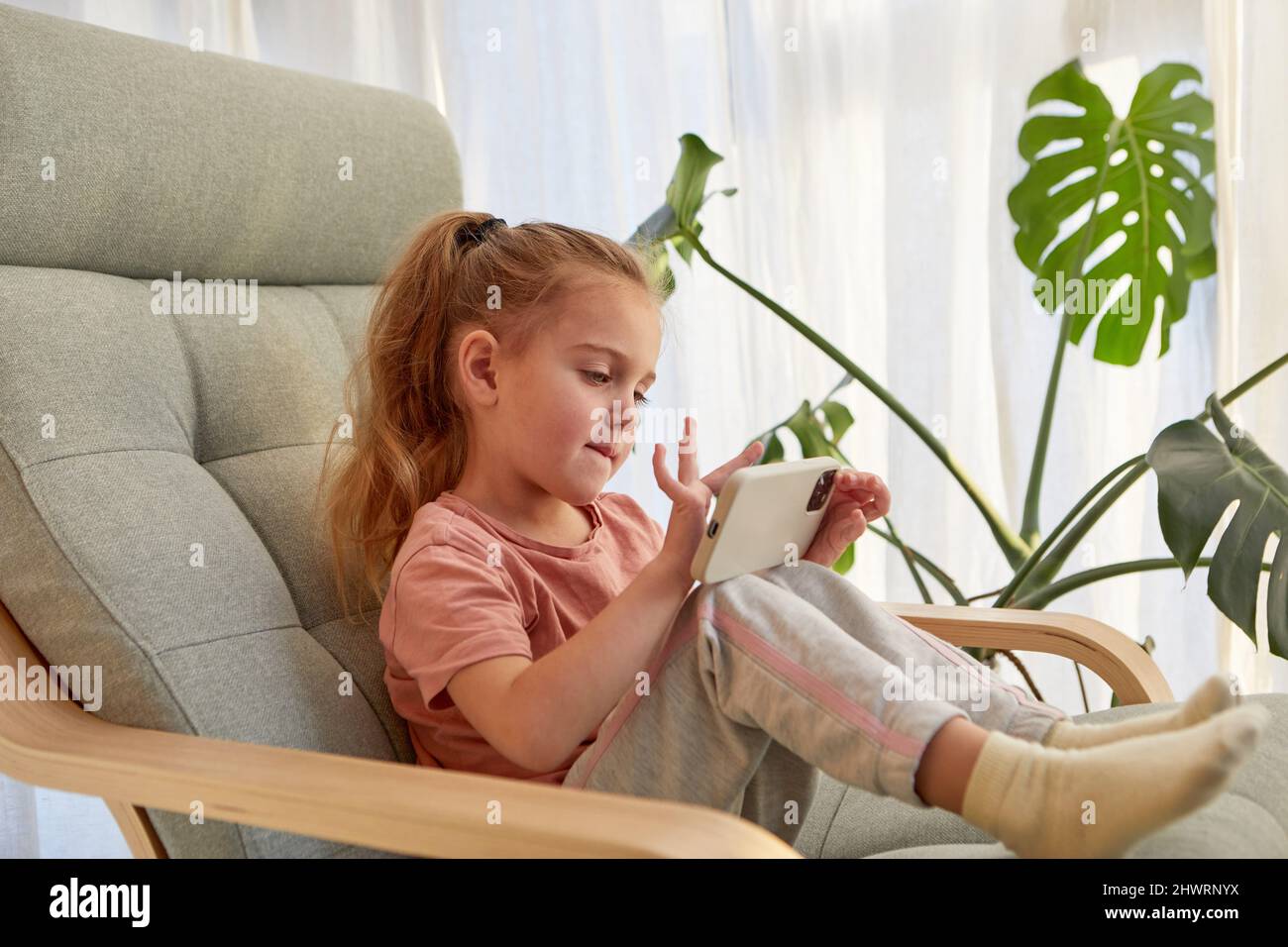 Vista lateral de linda niña pequeña con pelo largo rubio en ropa informal jugando a videojuegos en el teléfono móvil mientras se sienta en un cómodo sillón en dayli Foto de stock