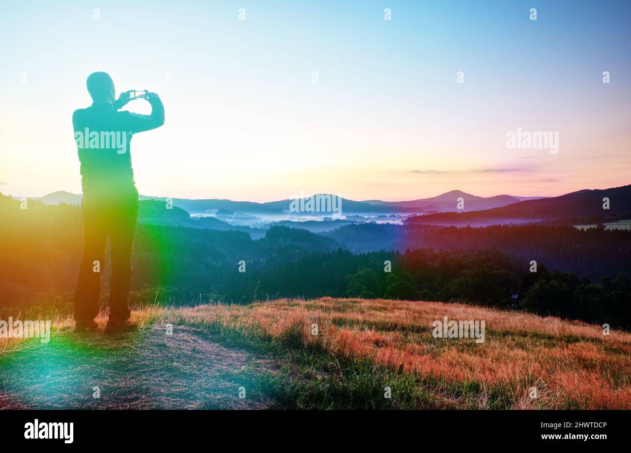 El hombre sostiene el teléfono por encima de la cabeza y toma fotos del paisaje brumoso de la mañana. Iluminación abstracta, llamaradas coloridas. Foto de stock