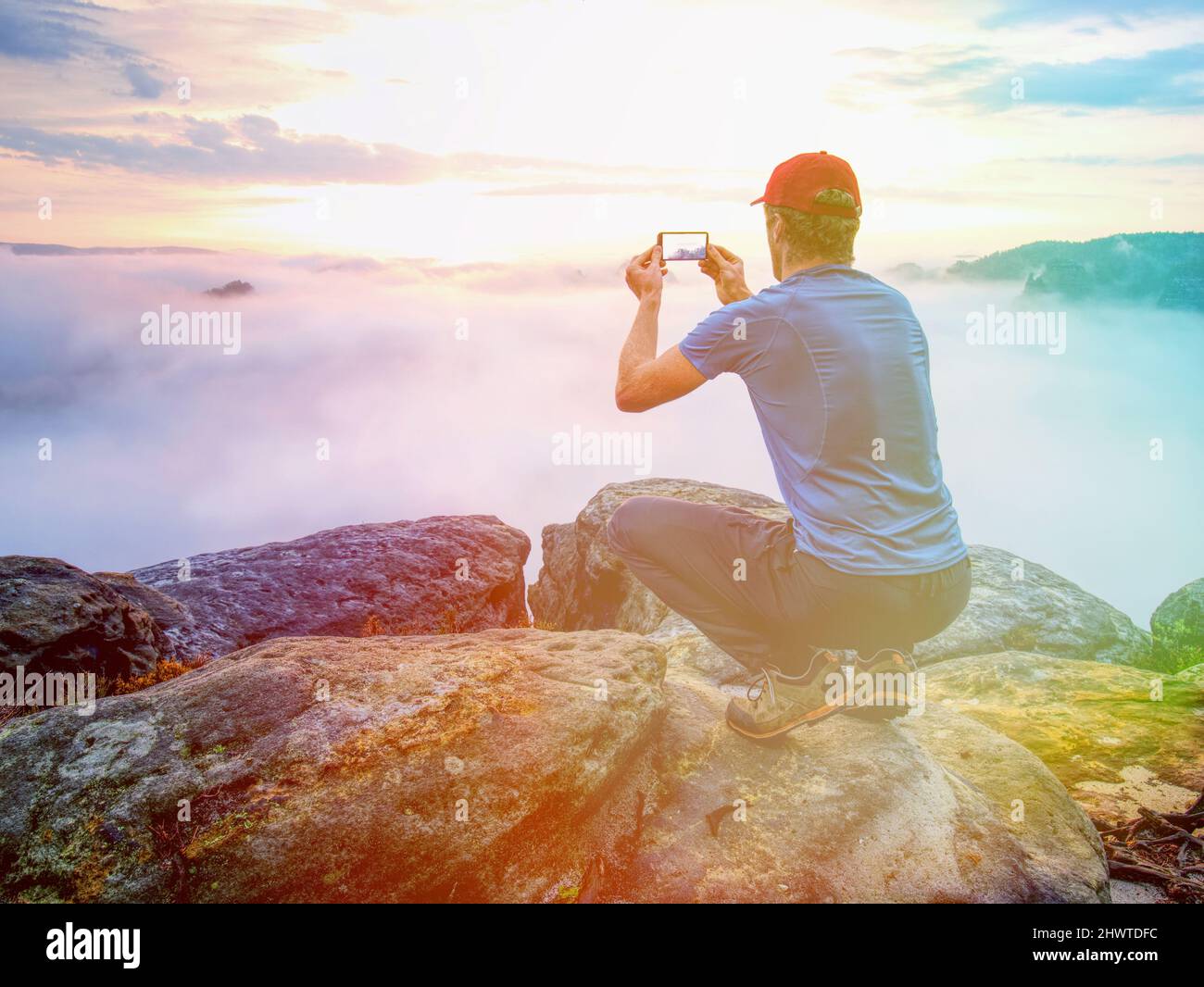 El hombre se sienta y toma una foto telefónica de la mañana malvada valle de montaña. Hiken en uso al aire libre. Iluminación abstracta, llamaradas coloridas. Foto de stock