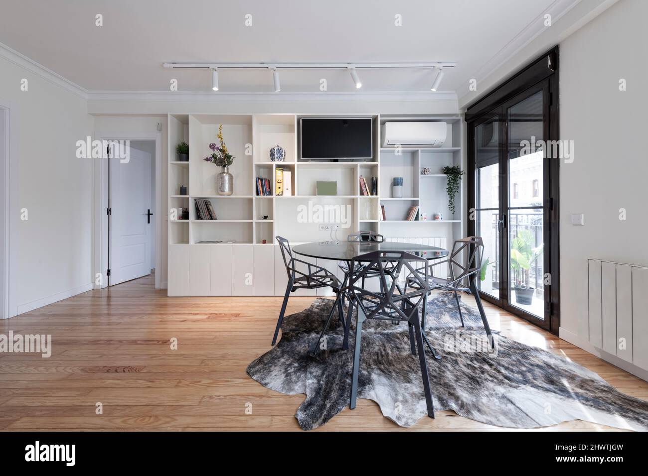 Sala de estar con mesa de comedor de cristal redonda sobre alfombra de  piel, sillas de diseño gris, ventana grande en una pared y estantería de  madera blanca Fotografía de stock -