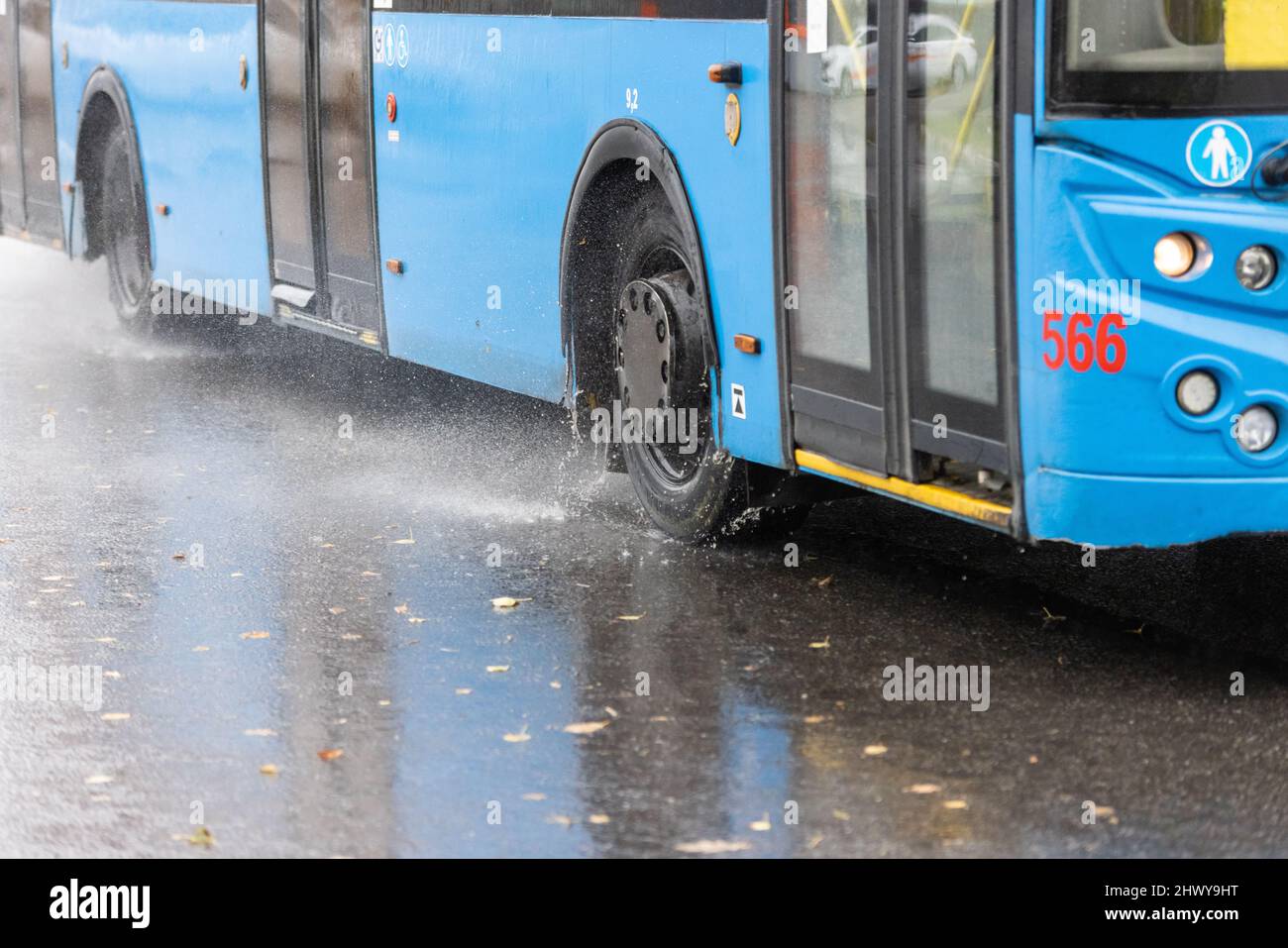 las salpicaduras de agua de lluvia fluyen desde las ruedas del trolebús azul moviéndose en la ciudad de luz del día con enfoque selectivo Foto de stock