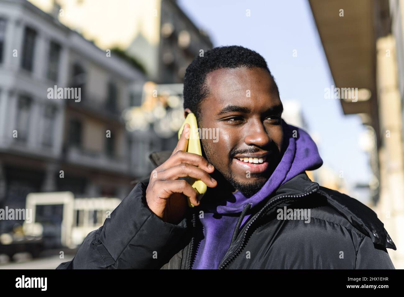 Alegre hombre afroamericano con capucha y ropa exterior sonriendo y mirando hacia fuera mientras responde a una llamada telefónica sobre un fondo borroso de la calle de la ciudad Foto de stock