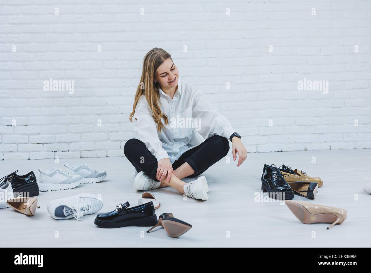 Una mujer joven se sienta en el suelo entre los zapatos y elige zapatos nuevos para sí Mujer en una camisa blanca y pantalón negro. Mujer con zapatos blancos Fotografía de