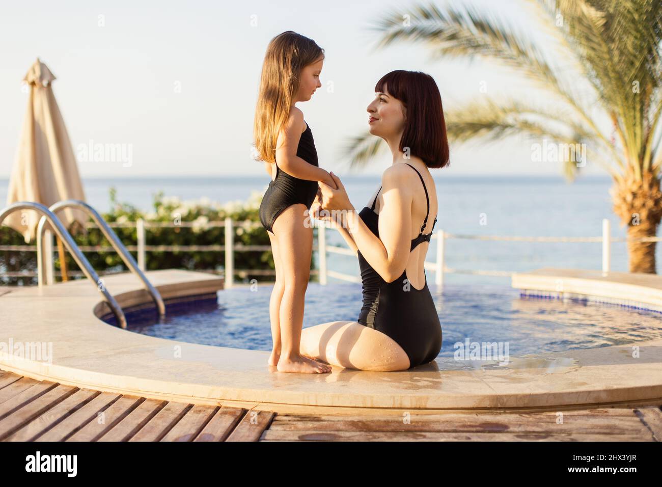 Madre e hija con bañadores negros sentados el borde de piscina en un hotel junto al mar. Se ven felices, de moda y ajustan sujetando las manos de los