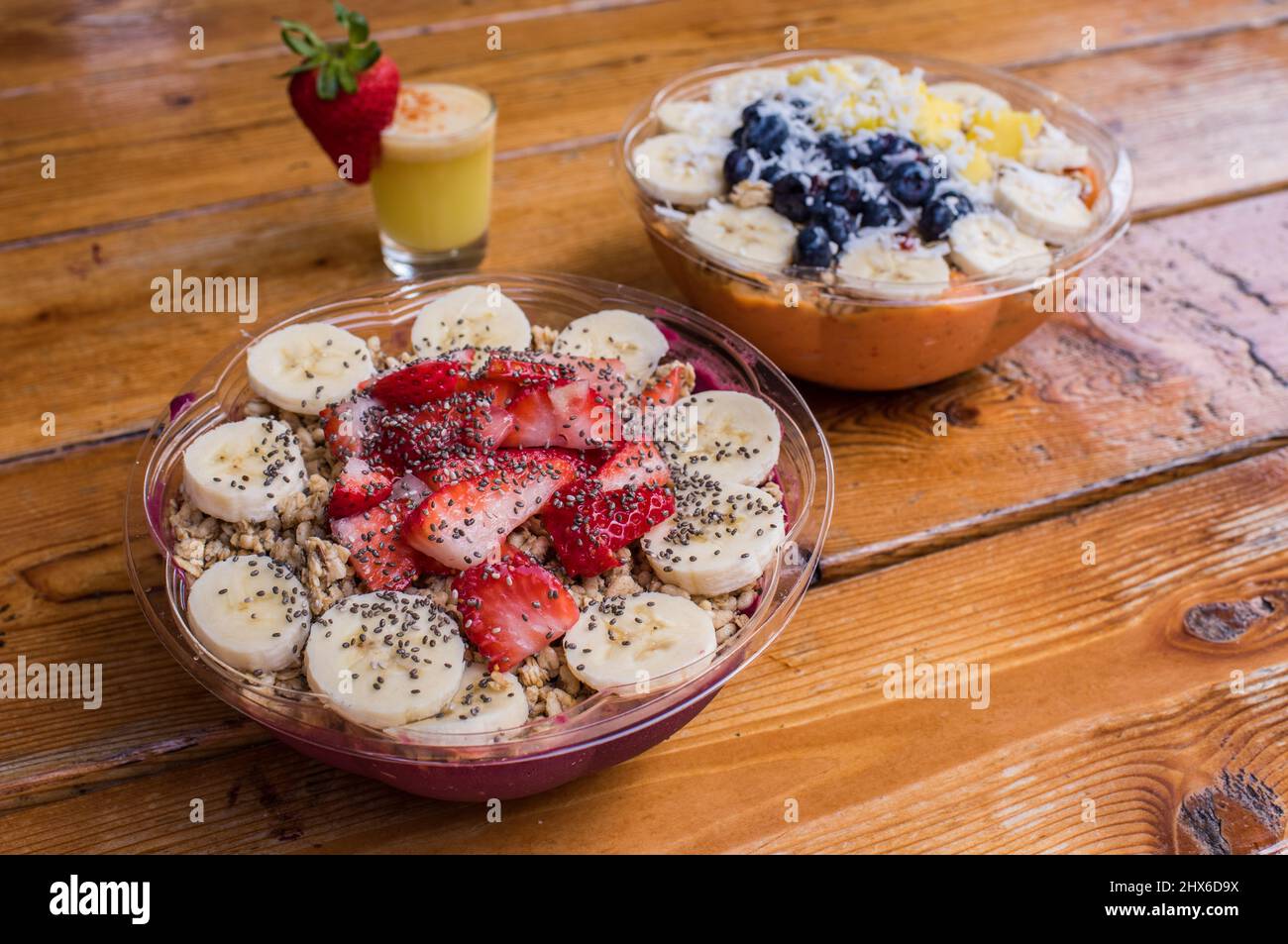 Dos cuencos de acai con bananas y fresas y un vaso de jugo con una guarnición de frutas en la mesa de madera de pino natural. Foto de stock