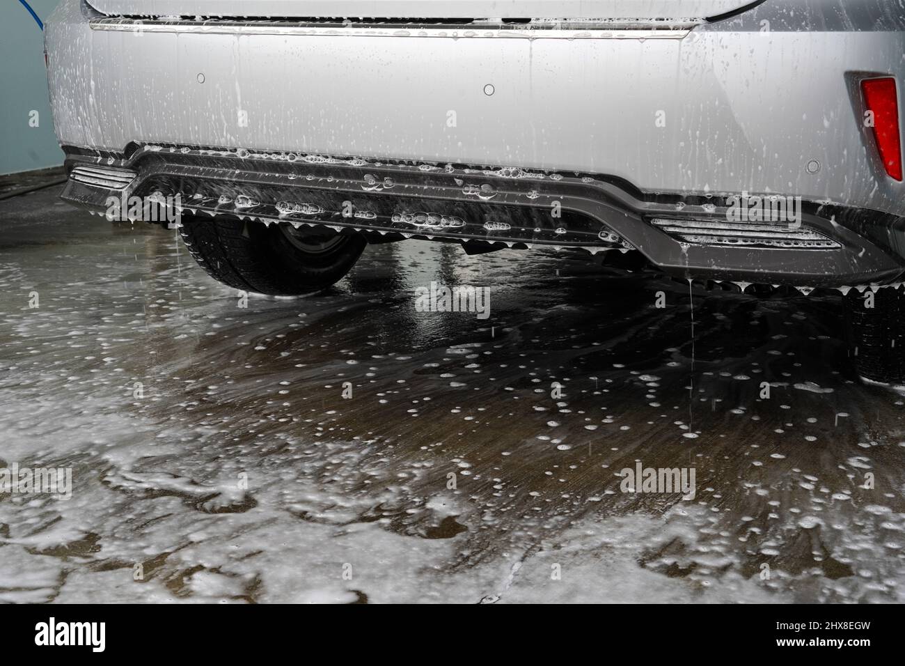 Gotea agua y espuma por el parachoques trasero de un coche después del lavado. El espacio de copia está en primer plano. La espuma y el agua se esparcen en el suelo. Foto de stock