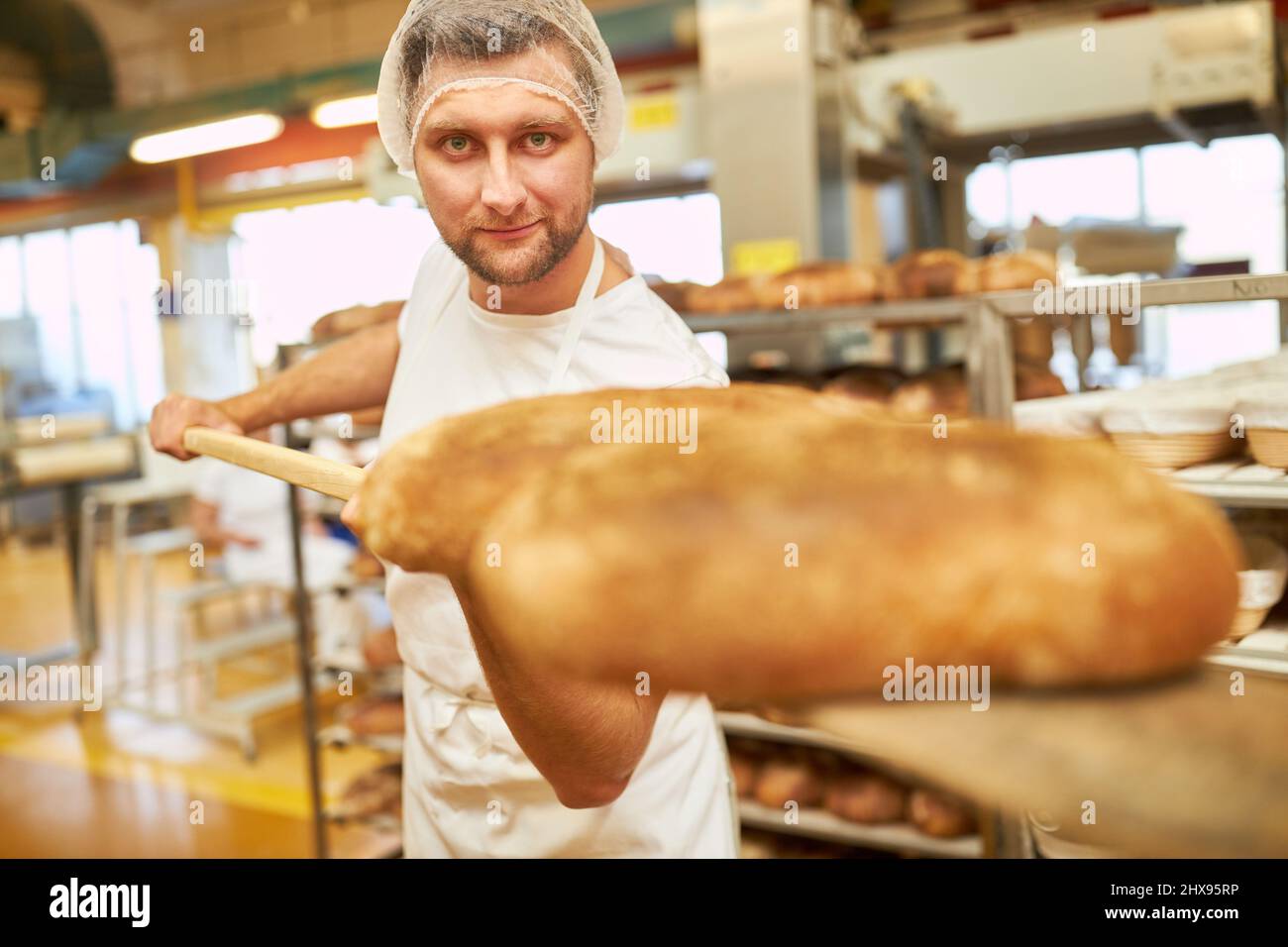 Aprendiz de Baker sosteniendo un empujador de pan con pan cocido en la habitación de atrás de la panadería Foto de stock