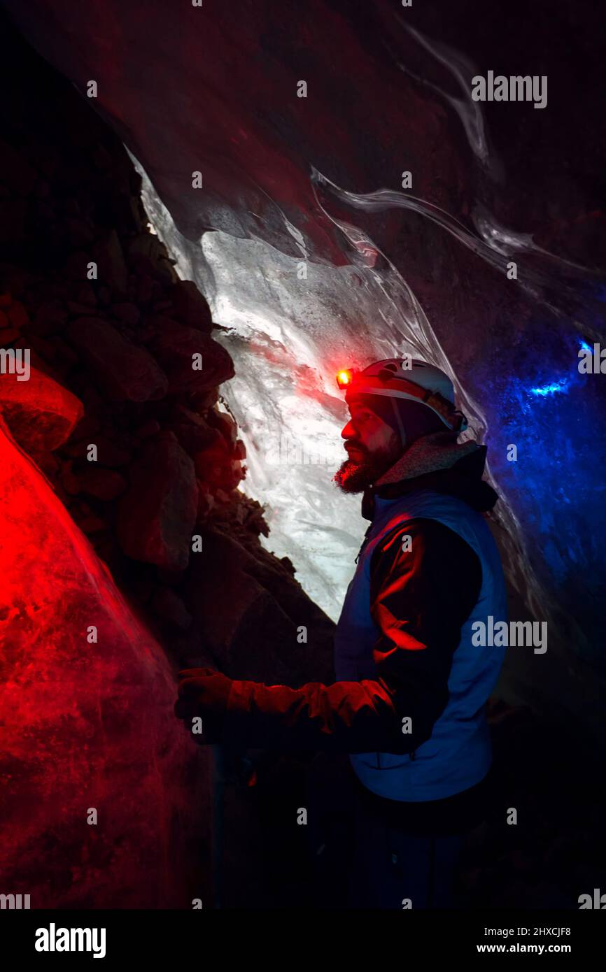 Alpinista con barba en casco con faro rojo brillante en profunda cueva de hielo glacial con luces azules explorar glaciar de montaña de invierno en Kazajstán Foto de stock
