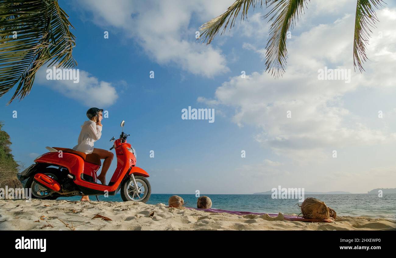 Viaje en scooter por carretera. Mujer sola en moto roja con ropa blanca en  la playa de arena junto al océano. Una niña caucásica paseo turístico cerca  de palmeras tropicales, el mar