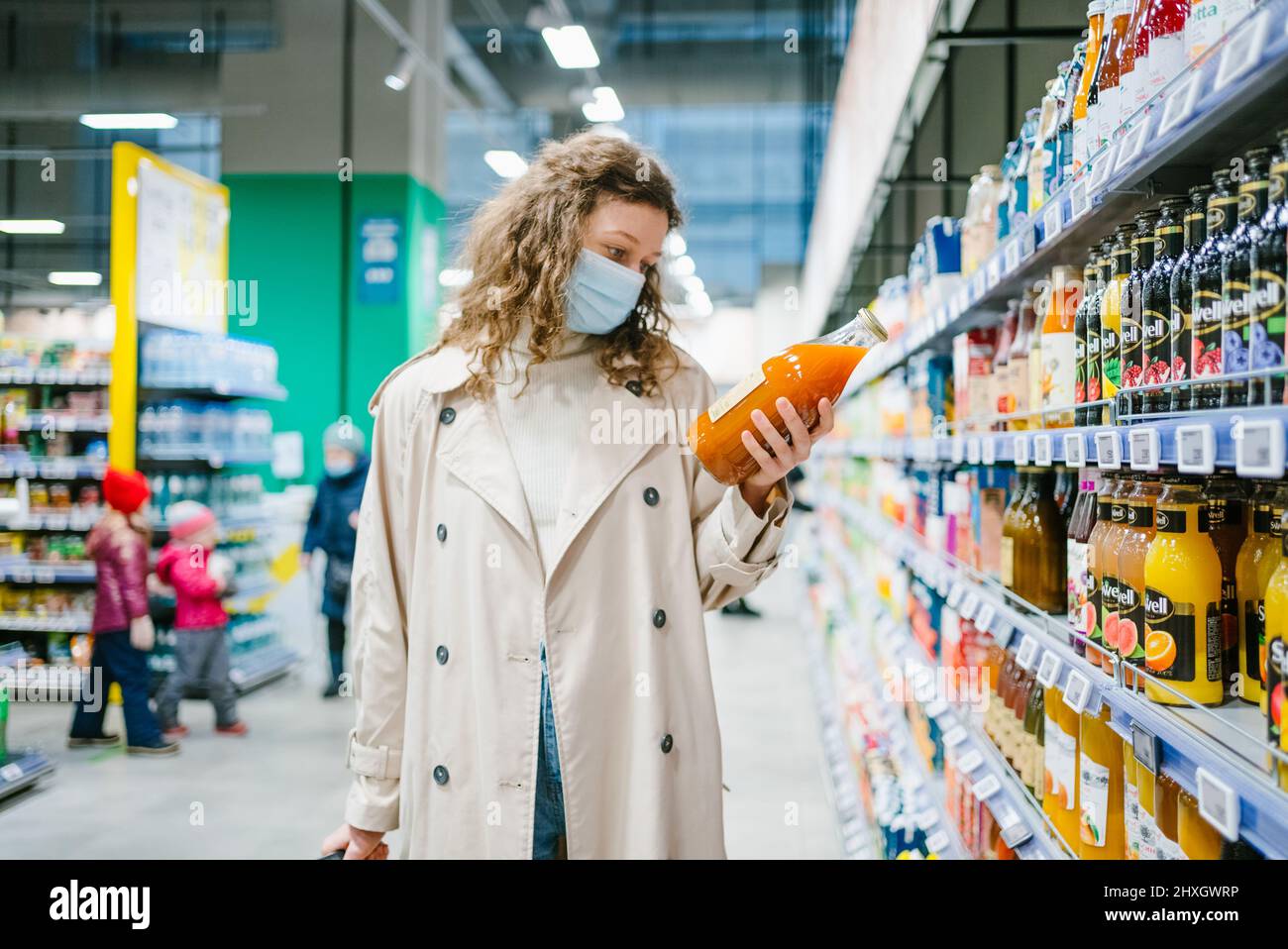 Una mujer rizado joven con una máscara médica y un impermeable beige elige el jugo en una botella de vidrio en un supermercado Foto de stock