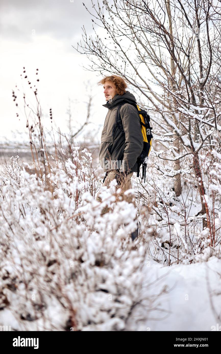 hombre con chaqueta abrigo y mochila camina por un camino cubierto de nieve hasta las montañas. Hermoso paisaje con nieve y bosque en el día nublado de invierno. Foto de stock