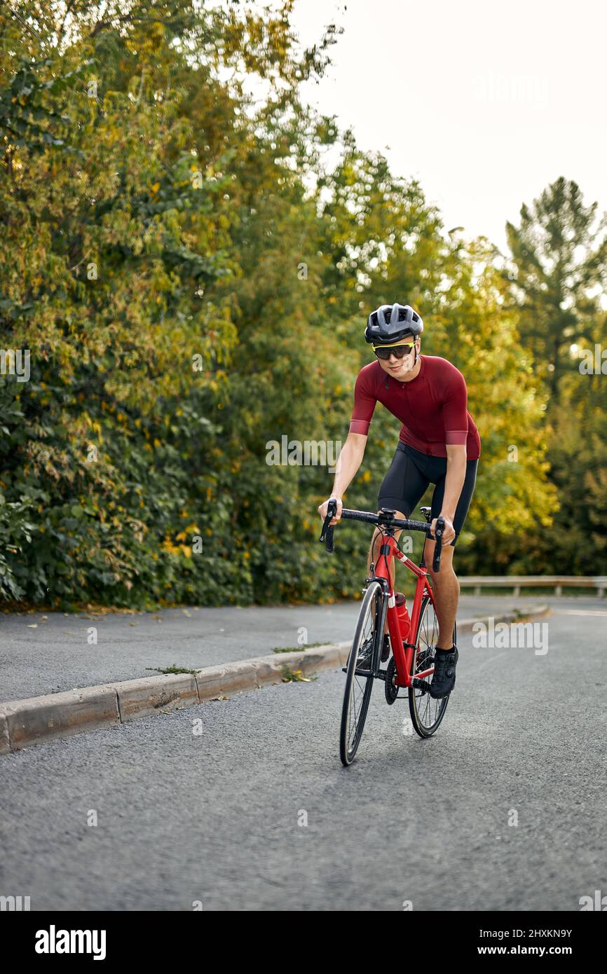 Hombre biker guapo en la ropa de ciclismo y casco de protección de entrenamiento en bicicleta, preparándose para la carrera en el parque al aire libre. Hombre disfrutando de la mañana bi Foto de stock