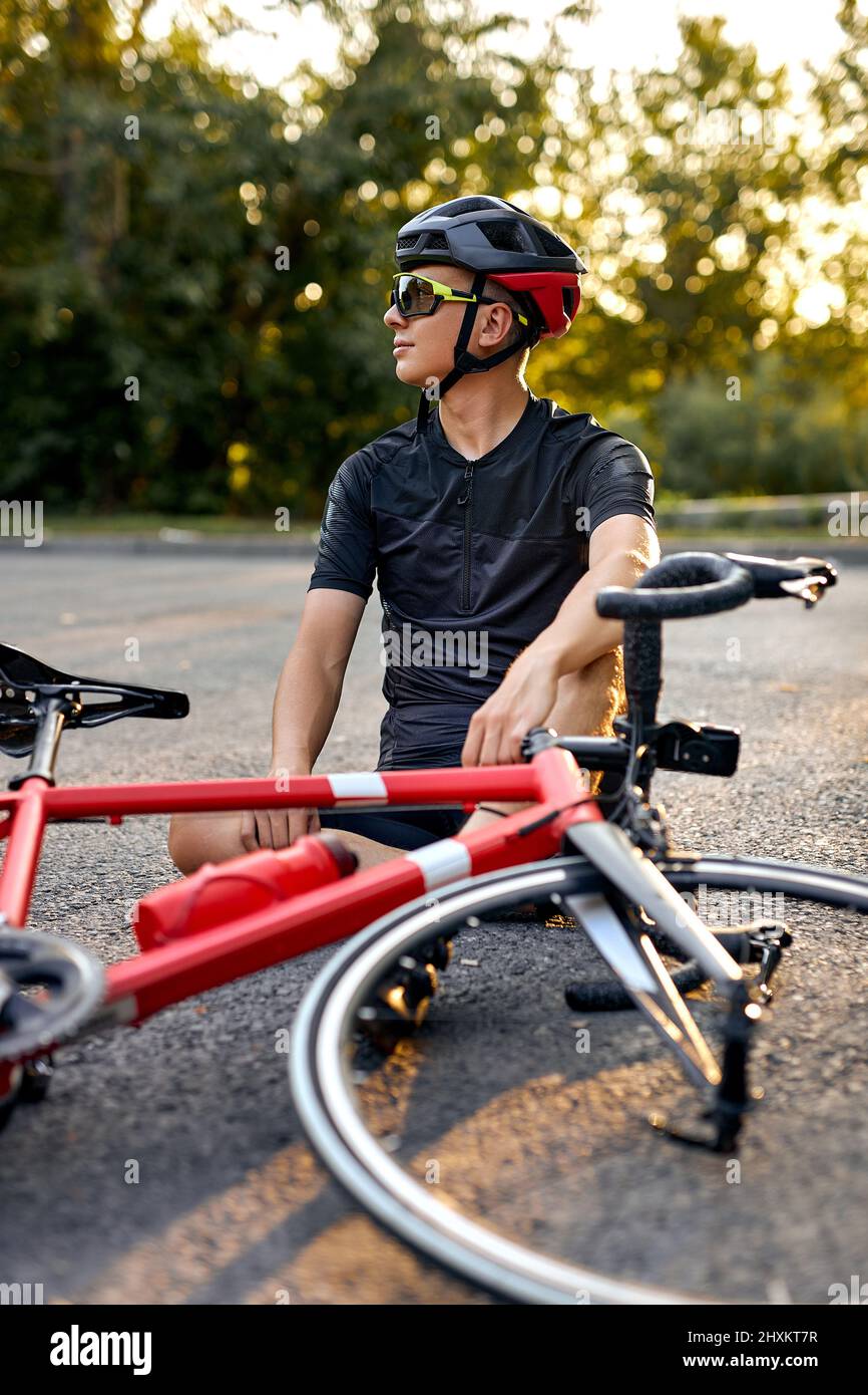 Ciclista tipo deportivo en bicicleta ropa y casco sentarse en la carretera cerca de la bicicleta, teniendo descanso. Hombre descansando y relajándose después de entrenar en bicicleta en la naturaleza. Él Foto de stock