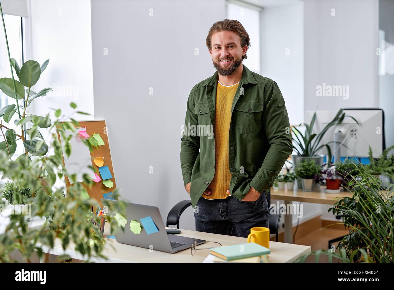 Retrato del sonriente diseñador de gerente caucásico en la oficina posando en la cámara, de pie detrás del escritorio con el portátil, muchas plantas en la sala de la oficina. Europeo Foto de stock