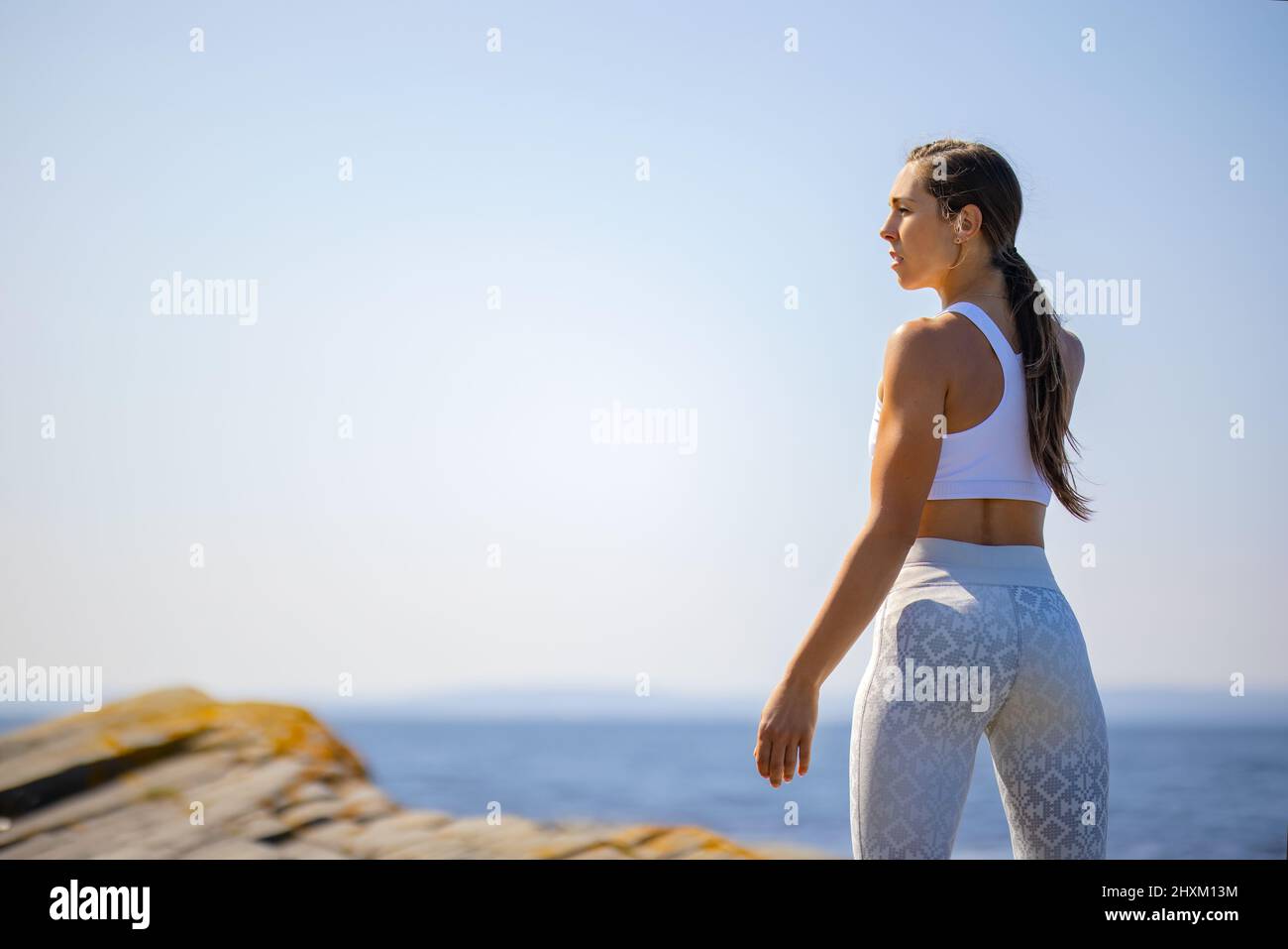 Vista trasera de una atleta descansando después de entrenar contra el cielo y el mar claros en un día soleado Foto de stock