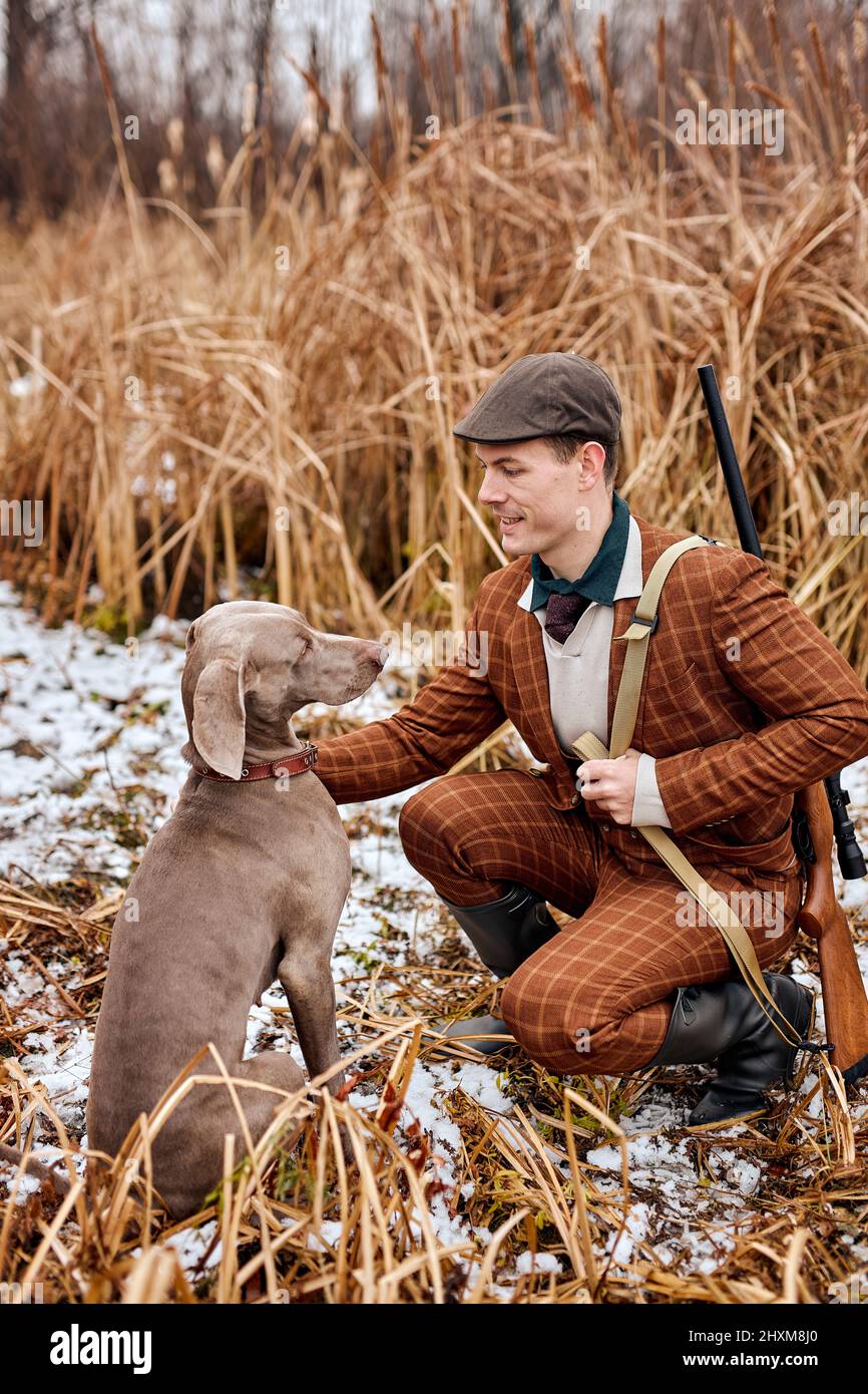 Joven cazador europeo caucásico en traje sentarse con el perro en el campo de partida para la caza, hombre guapo acariciando perro gris purebred y hablando, preparando Foto de stock