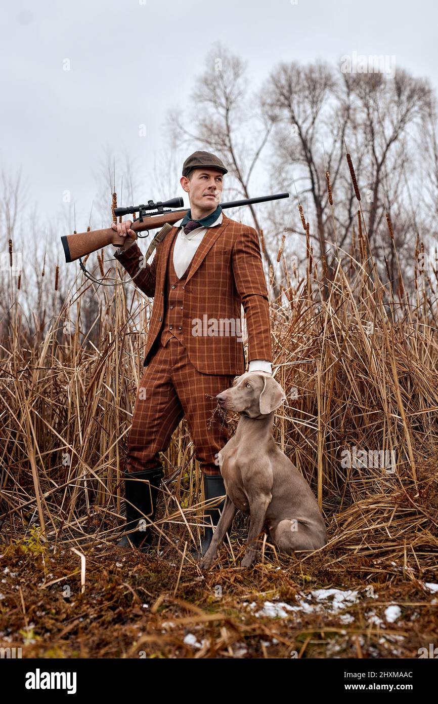 Período de caza, temporada de otoño abierto. Cazador caucásico joven con perro, arma en las manos en traje marrón de moda y sombrero en la naturaleza de la temporada de otoño, en s Foto de stock