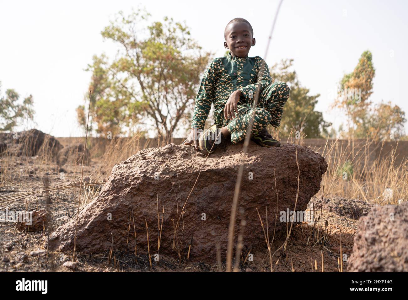 Lindo niño africano con una gran sonrisa sentado en una roca en medio de un páramo degradado y árido; problema ambiental Foto de stock