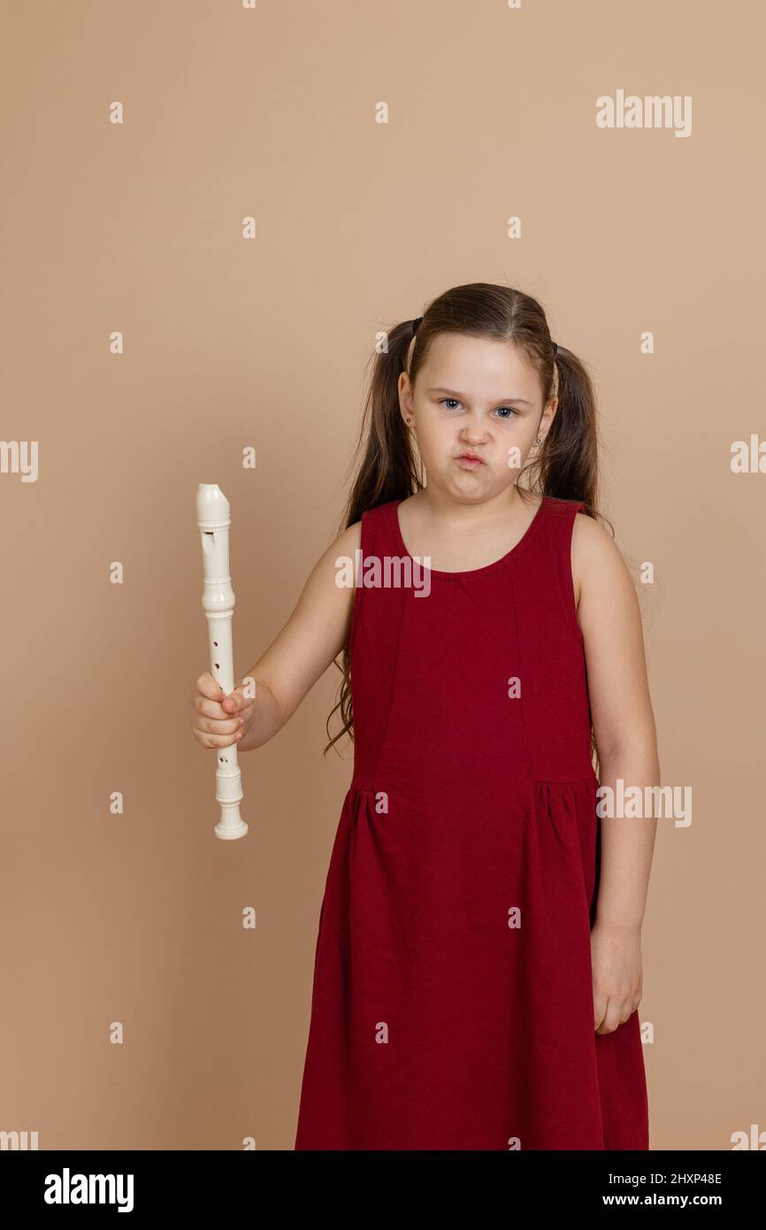 Chica de vestido rojo con expresión disgustada mantener la flauta común en la mano, fondo beige. Aprender a tocar instrumentos musicales de viento de madera. Foto de stock