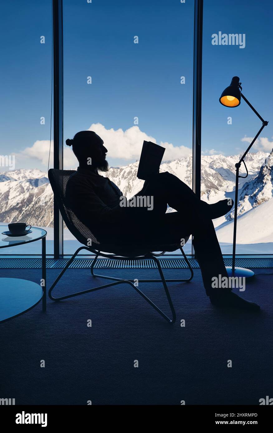 Hombre barbudo en silueta en la silla leyendo libro cerca de la ventana panorámica con vistas tranquilas de un hermoso paisaje nevado de montaña. Recubrir interior sm Foto de stock