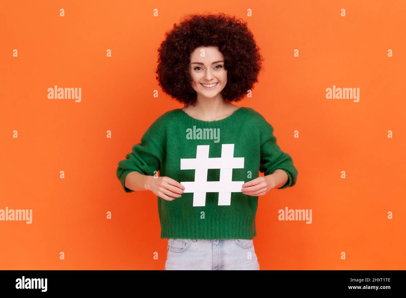 Mujer conmocionada con el peinado Afro apuntando al símbolo blanco hashtag, sorprendente promoción de tema viral en la red social, marcado de las tendencias del blog. Estudio en interior grabado aislado sobre fondo naranja. Foto de stock