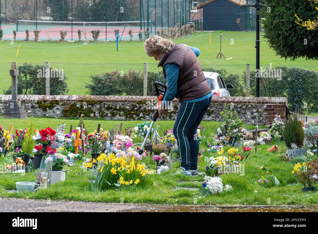 Mujer que corta hierba larga alrededor de las flores en un cementerio, timoriando para arriba, Hampshire, Inglaterra, Reino Unido Foto de stock