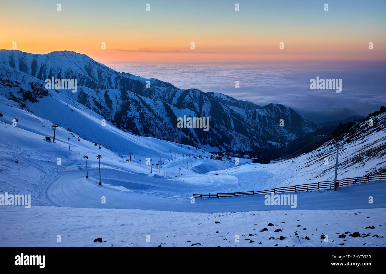 Hermoso paisaje Shymbulak estación de montaña nevada vacía pista de esquí y teleférico al atardecer cielo naranja en Almaty, Kazajstán. Foto de stock