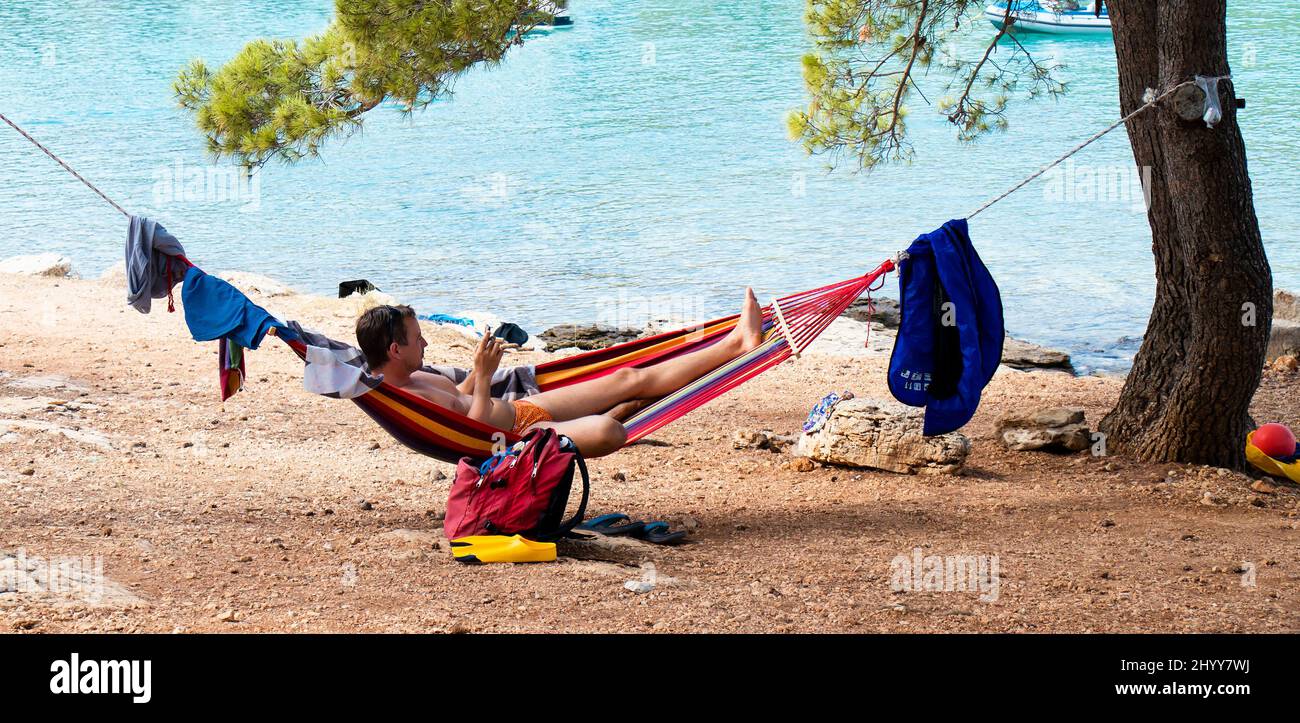 Persona tumbada sola en una colorida hamaca a la sombra de pinos en la playa en perfectas vacaciones de verano Foto de stock