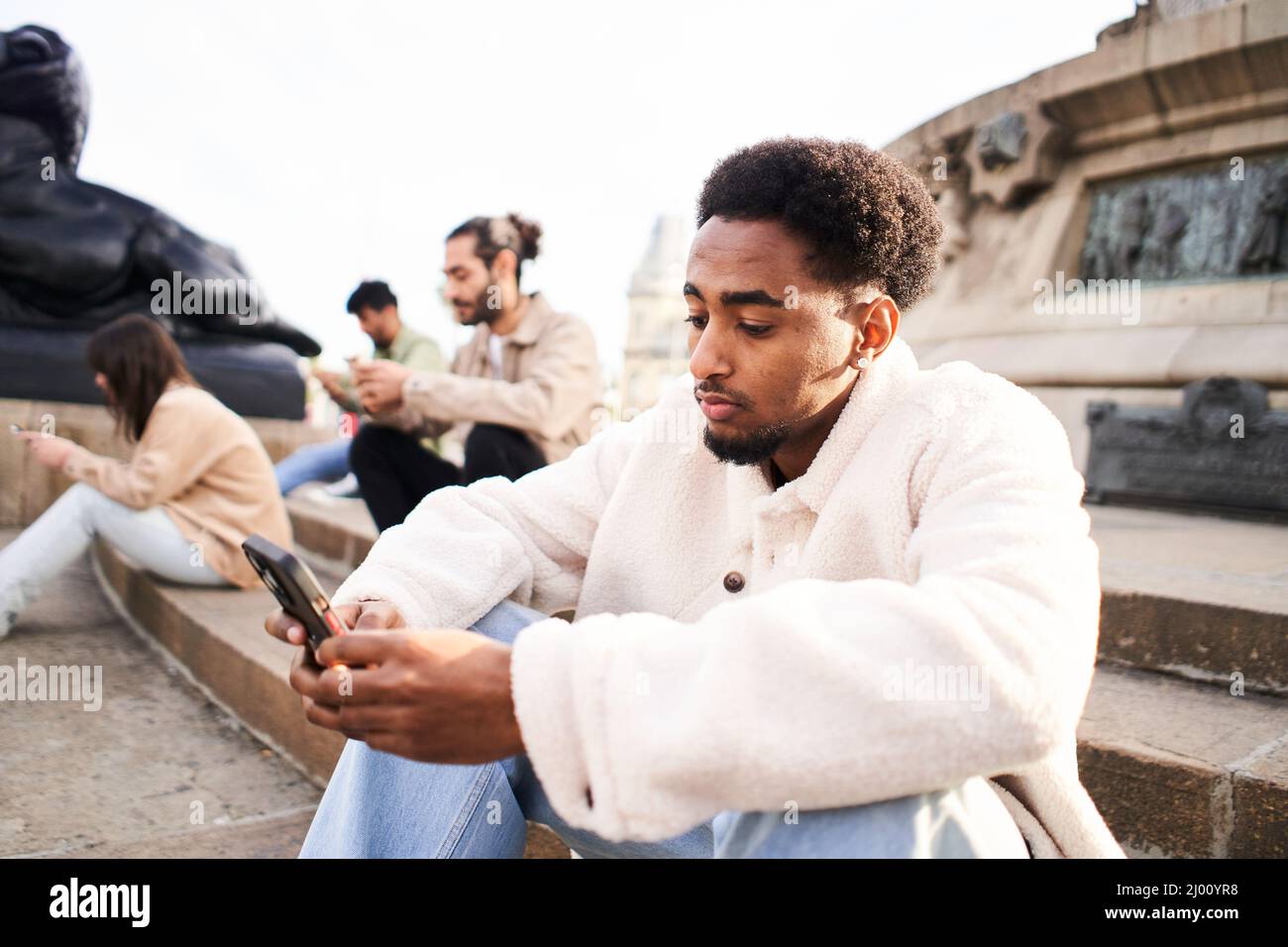 Un tipo afroamericano con célula móvil sentado en las escaleras de una ciudad con otros amigos ignorándose unos a otros mediante un smartphone. Grupo de adictos a la tecnología de Foto de stock