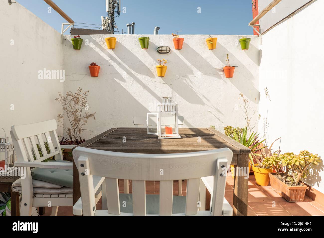 Pared blanca de una terraza con coloridas ollas colgantes de estilo andaluz,  una mesa de madera con sillas y más ollas en el suelo en un día soleado con  clo Fotografía de