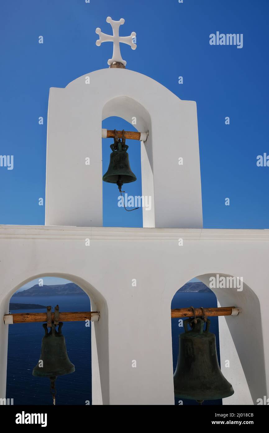 Tres campanas de la iglesia y una cruz religiosa en la parte superior junto a una iglesia y una hermosa vista del mar egeo en Santorini Foto de stock