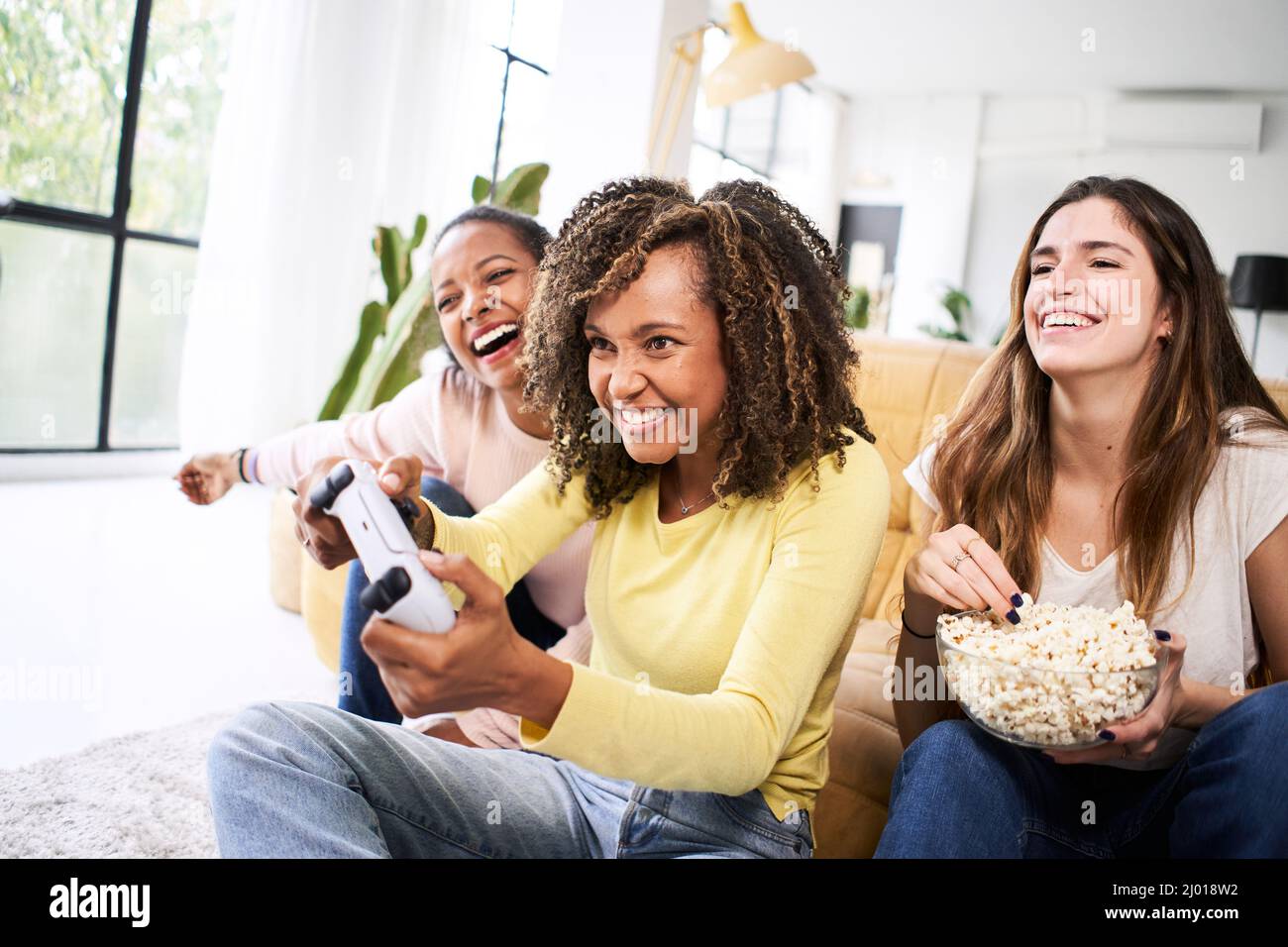 Competencia multirracial jóvenes amigos jugar juegos de vídeo juntos en casa. Sólo las mujeres jugando y comiendo papas. Foto de stock