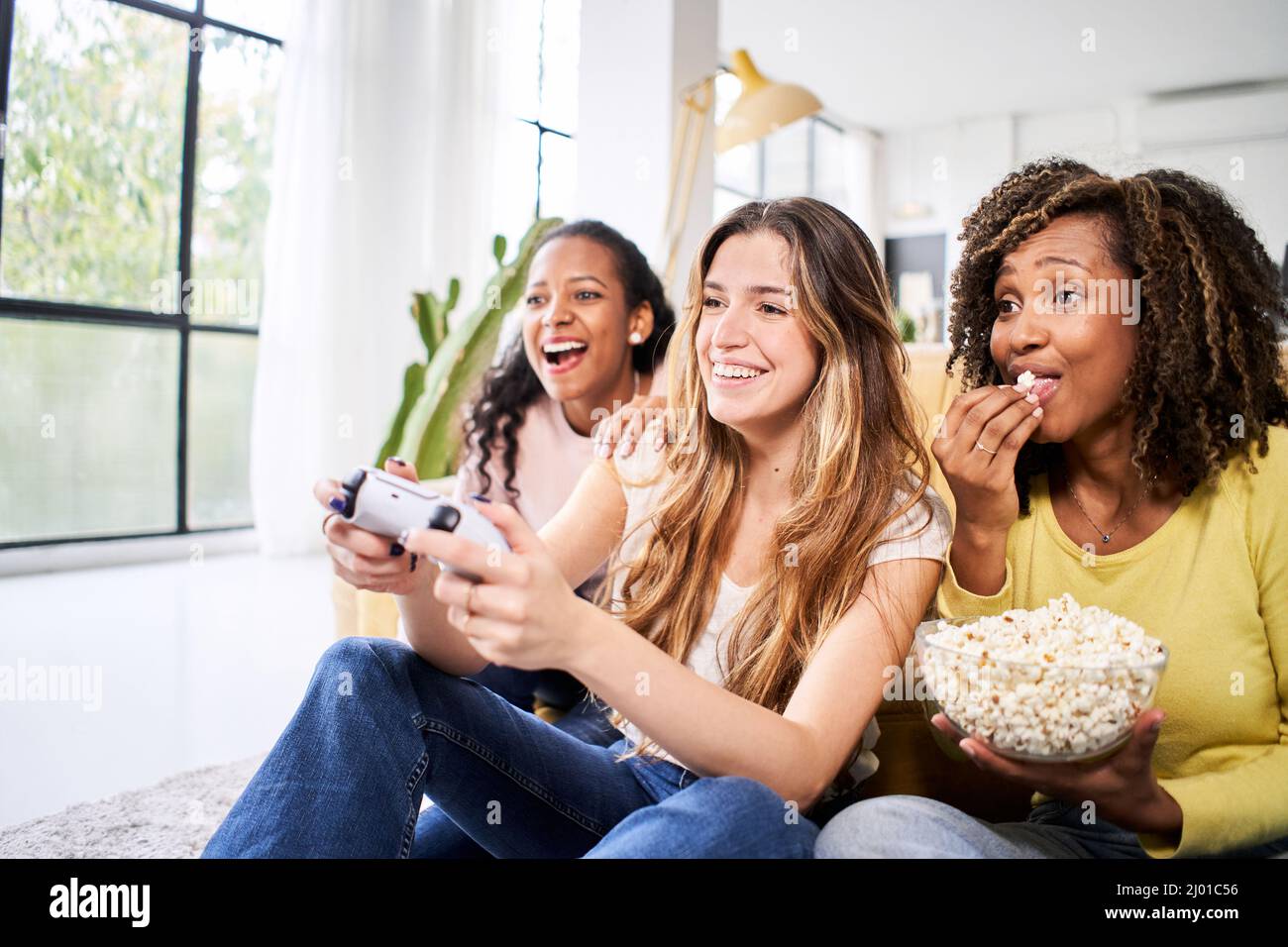 Grupo de jóvenes amigas multirraciales jóvenes juegan juegos de video juntos en casa. Sólo las mujeres jugando y comiendo papas. Foto de stock