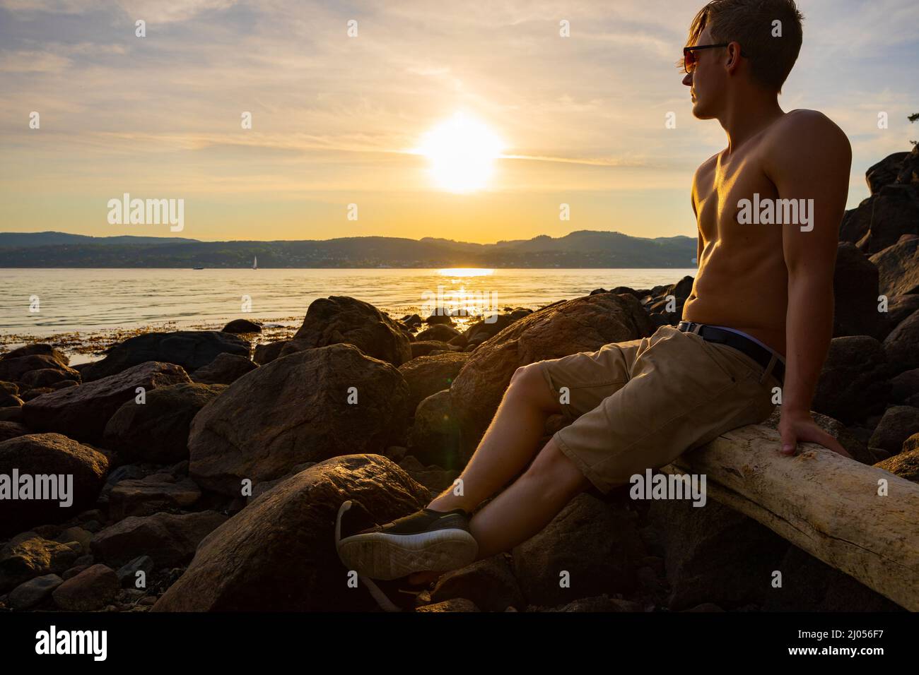 Hombre joven Shirtless sentado en la roca durante la puesta de sol Foto de stock