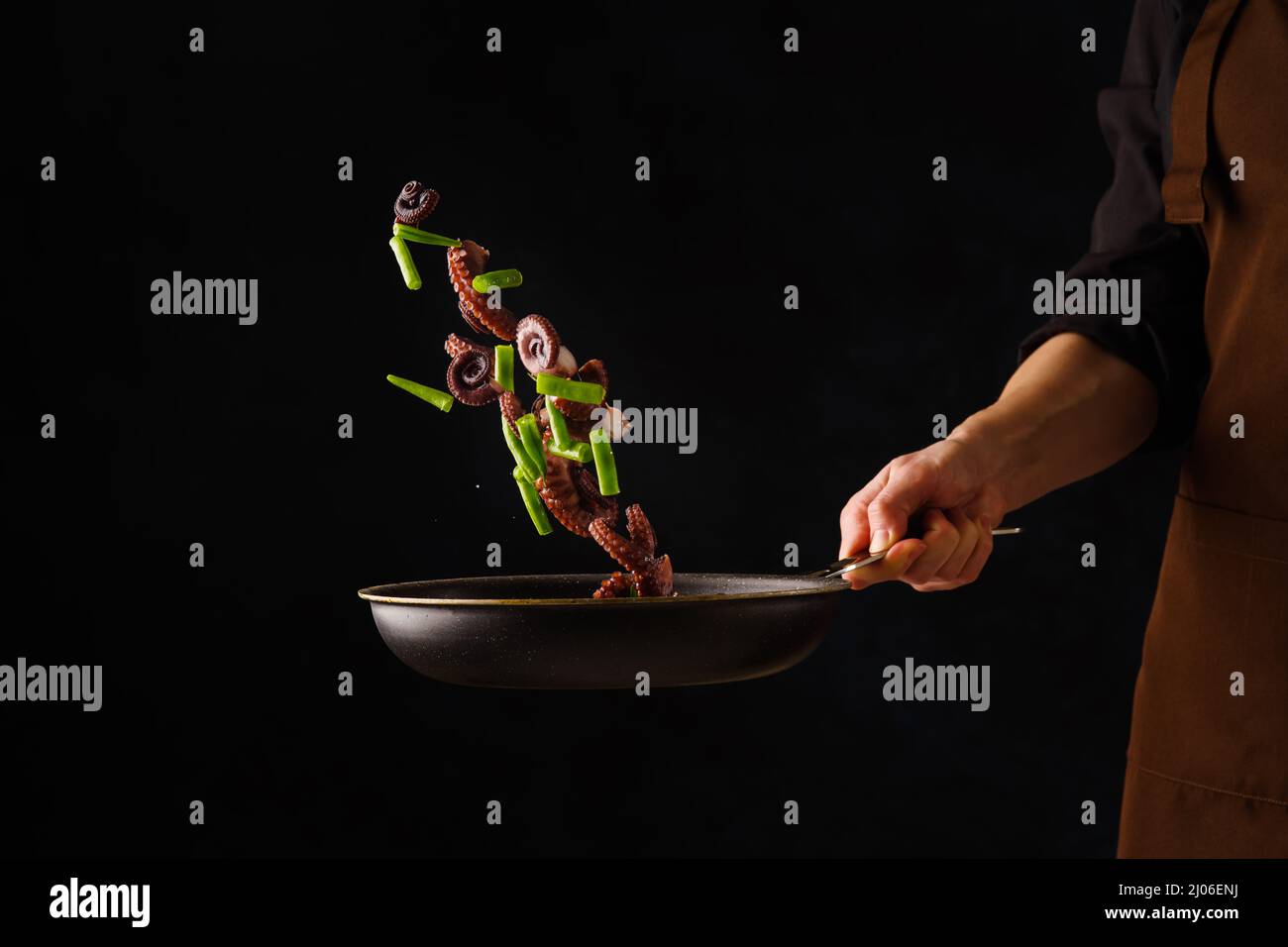 Mariscos - Pulpo con verduras en una sartén en un vuelo congelado en la mano del chef sobre un fondo negro. Minimalismo. Hay un espacio vacío para insertar. Foto de stock