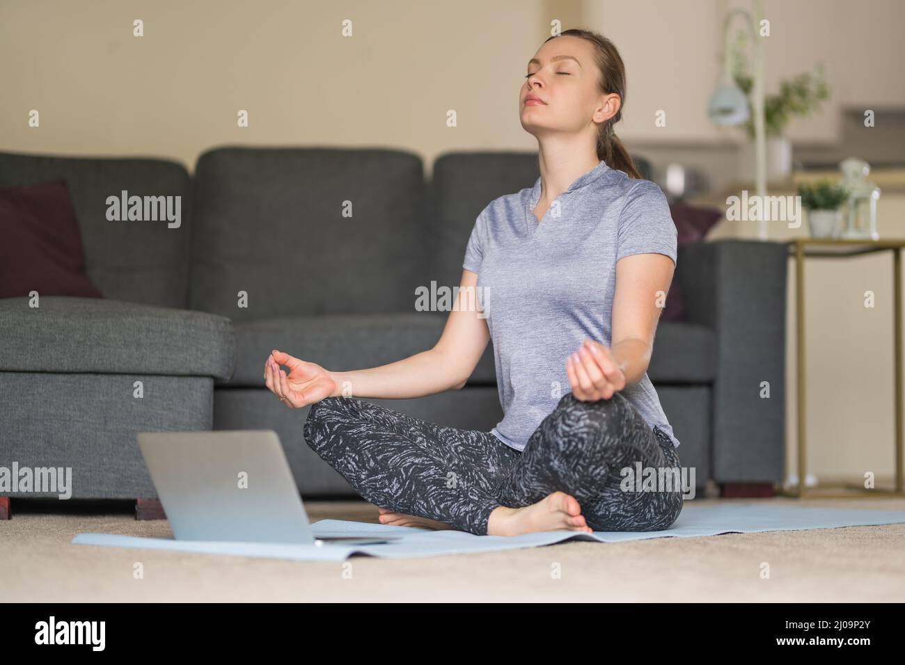 Una mujer joven en forma medita en la colchoneta y delante del ordenador portátil en la sala de estar de su casa. Concepto de relax y cuidado de la salud. Prácticas espirituales. Foto de stock