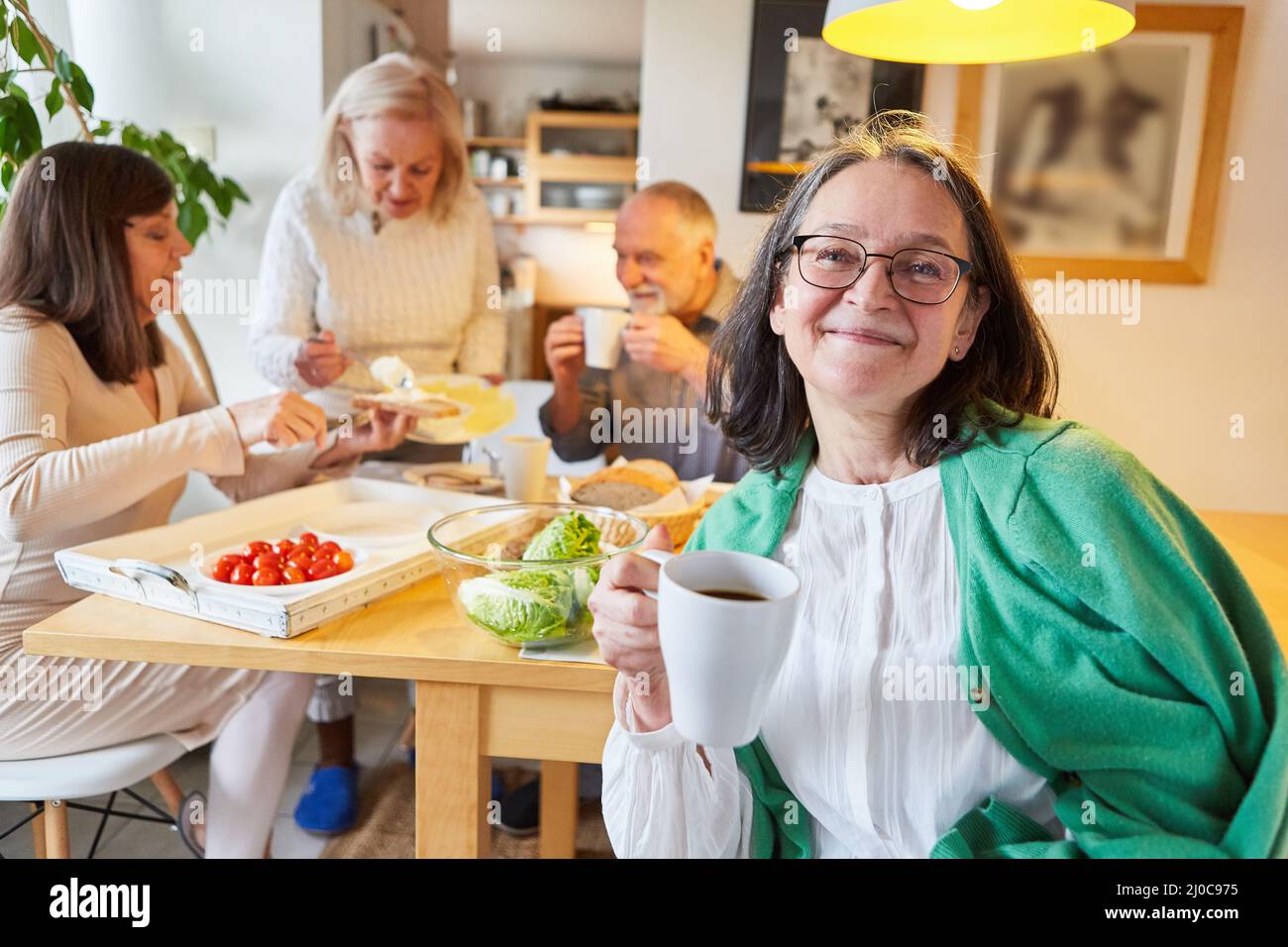 Mujer mayor tomando una taza de café en la cocina con amigos desayunando en el fondo Foto de stock