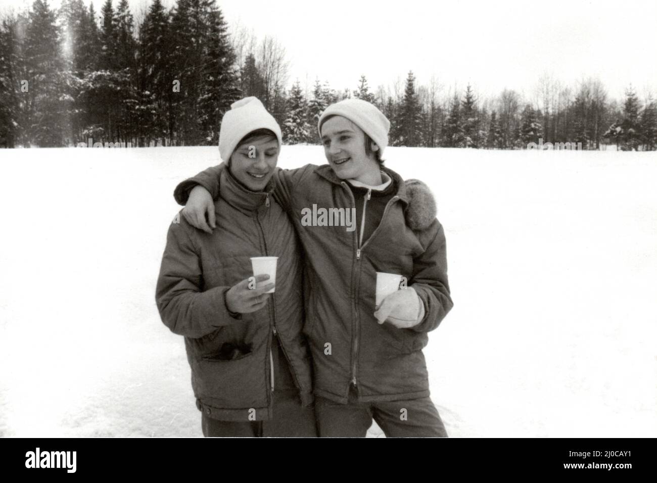 Auténtica fotografía vintage de dos sonrientes chicos adolescentes felices bebiendo de vasos de plástico de pie en la nieve cerca del bosque, Suecia, concepto de felicidad Foto de stock